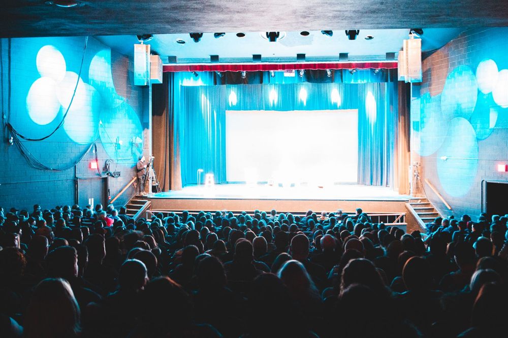Packed audience facing a brightly lit theater stage